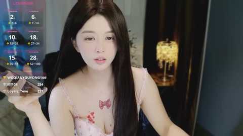 Video of a young Asian woman with long black hair, wearing a pink lace bra, seated in a dimly lit room.