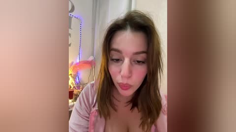 Video of a young Caucasian woman with shoulder-length brown hair, wearing a pink top, making a kissy face. Background features a room with a pink unicorn toy, white walls, and a colorful LED strip.