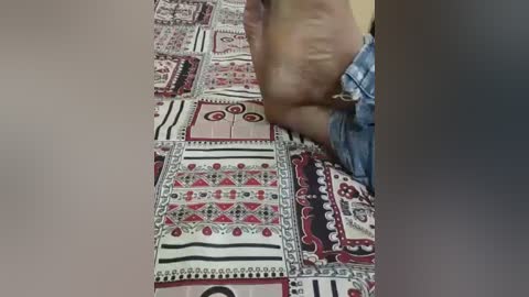 Media: Video of a person's head and shoulders, partially cropped, wearing a blue denim shirt, resting on a colorful, patterned quilt with intricate designs in red, black, and white.