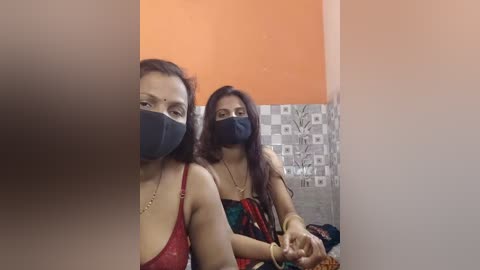 Video of two South Asian women in a bathroom with orange walls; one wears a red bra, the other a colorful saree, both masked, holding a phone.