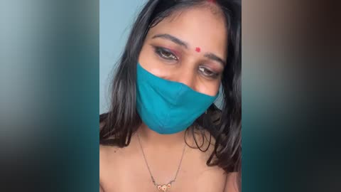 Video of a South Asian woman with medium skin tone, long black hair, wearing a turquoise mask, red bindi, and a gold necklace.