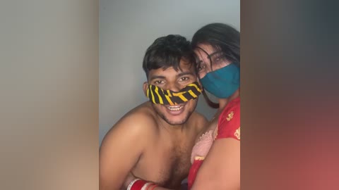 Video of two smiling South Asian men, one topless with a yellow and black bandana, and the other in a red shirt, both wearing blue masks, in a dimly lit room.
