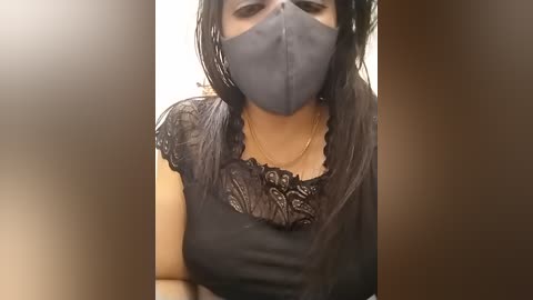 A video of a woman with medium skin tone, wearing a black lace top, black face mask, and long black hair. She stands in a dimly lit room with blurred, brownish tones.