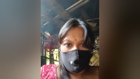 Video of a woman in a traditional red and yellow saree, wearing a black mask, with a red dot on her forehead. Background shows a train compartment with dim lighting and another person in the background.