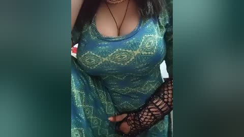 Video of a woman with medium brown skin, wearing a green paisley-patterned dress with a low-cut neckline revealing ample cleavage. She has a black fishnet glove on her right hand, and her long dark hair falls over her shoulders.