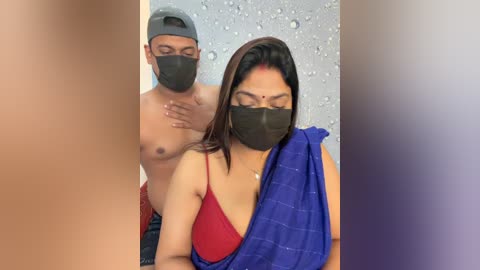 Video of a South Asian man in a gray cap, mask, and red shirt standing behind a woman with long dark hair, wearing a blue saree and red bra, both indoors.