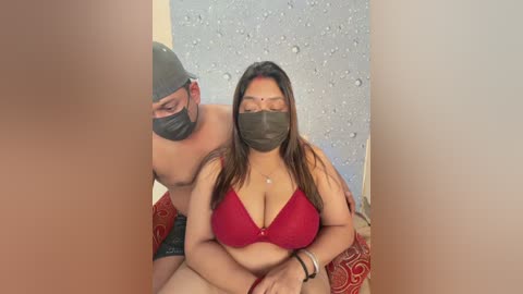 A video of a woman with medium skin tone and long hair in a red bra, wearing a face mask, sitting between two men, one with a beard, in a dimly lit room with a textured wall.