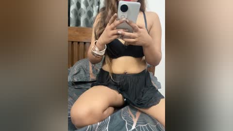 Video of a woman with long, wavy brown hair, wearing a black bra and shorts, sitting on a bed with a patterned blanket. She holds a smartphone, partially covering her face.