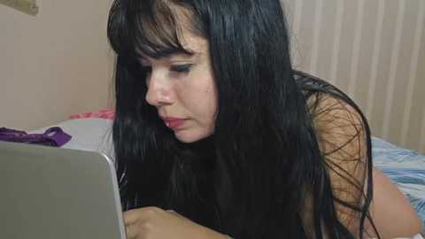 Media: Video of a young woman with long black hair and pale skin, wearing a sheer top, intensely focused on a laptop in a dimly lit room with beige walls and a bed.