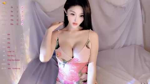 A video of an East Asian woman with long, straight black hair, wearing a low-cut floral dress, posing on a bed with white sheets. The background includes a white curtain and a digital calendar displaying dates.