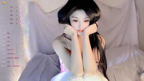 A video of a young Asian woman with long, straight black hair, wearing a white lace top, lying on a bed with soft, white sheets. The background features a draped curtain and a calendar overlay with social media icons.