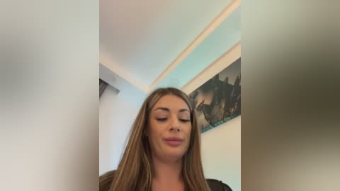 Video of a young woman with long, straight, light brown hair, wearing a black top, standing in a room with light blue walls. Behind her is a large, abstract painting.