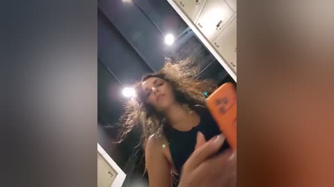Video of a young woman with long, curly hair taking a selfie in a modern, dimly-lit room. She wears a black tank top and holds an orange phone. Background includes metallic cabinets and recessed lighting.