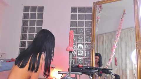 Video of a nude woman with long black hair, standing in front of a mirror, surrounded by pink and white floral decorations, and a pink dildo on a chair.
