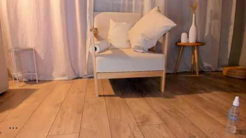 Video of a minimalist room with a white, cushioned armchair, white pillows, a small wooden side table, white sheer curtains, and wooden floor.
