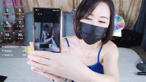 A video of an East Asian woman with short black hair, wearing a black face mask, a blue tank top, and a cross necklace, holding a smartphone showing a man's face, in a room with a fan and a wall mirror.
