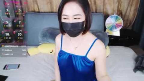 Video of an East Asian woman with short black hair, wearing a blue satin camisole, black face mask, and sitting on a bed with a yellow teddy bear and colorful spinning toy.