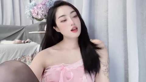 Video of a fair-skinned, young Asian woman with long black hair, wearing a pink lace top, smiling seductively, in a softly lit room with light curtains and a floral arrangement.
