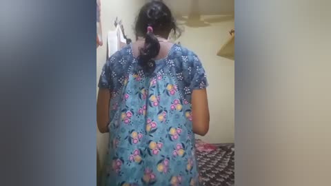 Video of a woman with medium brown skin, wearing a blue floral dress, standing in a dimly lit room with a bed covered in a patterned blanket.