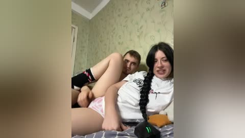 Video of a young couple in a bedroom, the woman with long black hair and braids, wearing a white shirt, lying on a bed, and the man with short brown hair, wearing black socks, engaging in sexual activity.