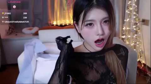 Video of a young Asian woman with long black hair, wearing a black lace top, in a cozy, warmly lit bedroom with fairy lights, bed, and a black cat on her shoulder.