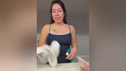 A video of a young woman with long brown hair, wearing a dark blue tank top and white gloves, cleaning a white countertop with a green sponge.