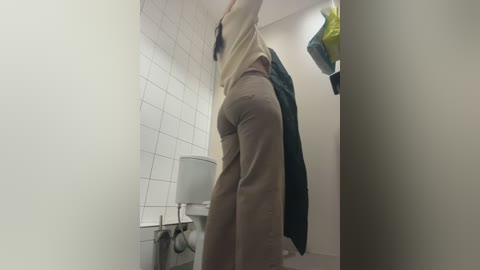 Video of a woman in a bathroom, wearing a beige shirt and pants, standing near a urinal and white-tiled wall.