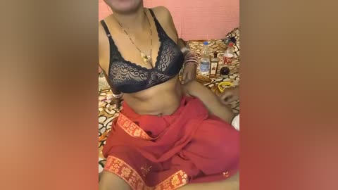 Video of a topless woman with medium brown skin, wearing a black lace bra and red sari, sitting on a bed with a floral patterned blanket.