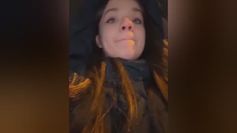 Video of a young woman with fair skin and brown hair, wearing a black coat, smiling. The image is slightly blurry, with a warm, golden hue in the background.