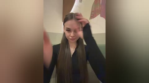 Video of a young woman with long, straight brown hair, wearing a black long-sleeved shirt, standing in a bathroom with a blurred reflection of herself and a partially visible man.