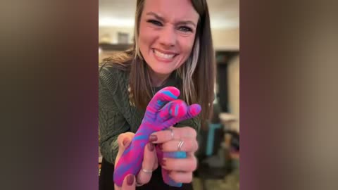 Video of a smiling woman with shoulder-length brown hair, wearing a green sweater, holding a pair of vibrant pink and blue striped socks. The background is blurred, showing a homey interior.