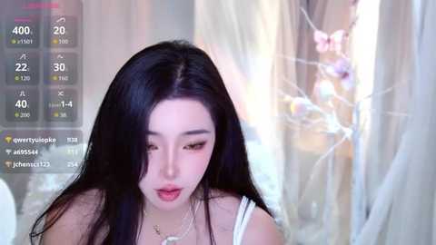 Video of an East Asian woman with long black hair, wearing a white dress, looking down. She has light skin and is in a softly lit room with sheer curtains.
