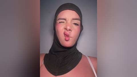 Video of a young woman with light skin, wearing a black hijab and making a playful face with puckered lips. She has on a white spaghetti strap top, set against a plain grey background.