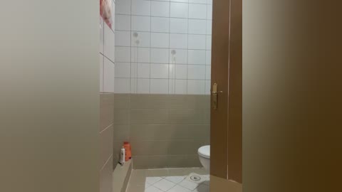 Video of a minimalist bathroom with white tiled walls and floor, a wooden door, a toilet, and a small orange bottle on the left shelf.
