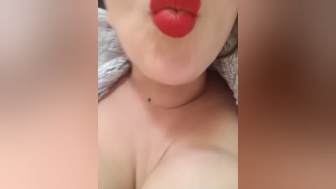 Video of a close-up of a fair-skinned woman's face with full red lips, kissing her own neck. Her skin is smooth, and she's wearing a gray, textured cardigan. The background is blurry.