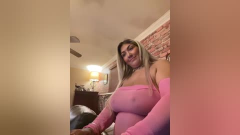 A video shows a curvy, blonde woman with long hair in a pink off-shoulder top, seated in a cozy living room with a brick wall and warm lighting.