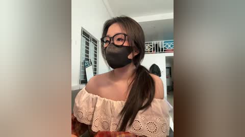 Video of a young woman with medium skin tone, long dark hair, wearing black-rimmed glasses, a black mask, and a beige off-shoulder lace top, indoors near a window with a blue and white pattern.