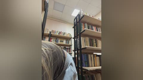 Video of a library interior, showing a person with long blonde hair, wearing a light blue shirt, bent over a bookshelf. The shelves are filled with books, and the room has a white ceiling with fluorescent lights.