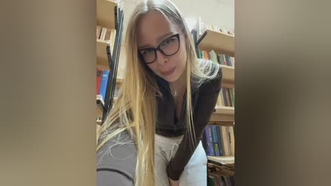 Video of a blonde woman with long hair, wearing black-framed glasses and a dark top, leaning forward in a library with wooden bookshelves filled with books.