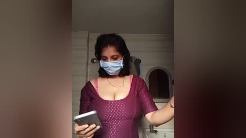 A video shows a woman with medium skin tone, wearing a blue surgical mask, a purple sequined top, and a gold necklace. She is holding a smartphone, standing in a dimly lit bathroom with beige walls.