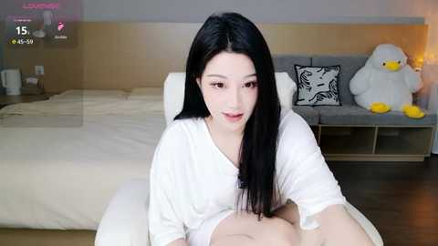 A video of an Asian woman with long black hair, wearing a white shirt, sitting on a white couch in a modern bedroom with a gray bed, a stuffed duck, and a digital clock.