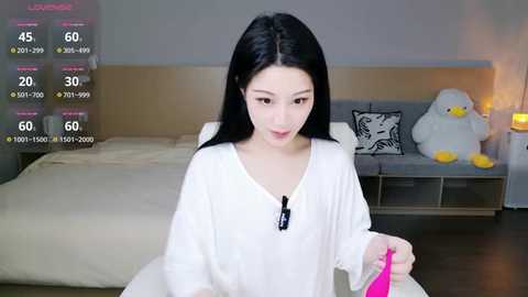 Video of an East Asian woman with long black hair, wearing a white V-neck top, sitting in a modern bedroom with a gray sofa and plush toys.