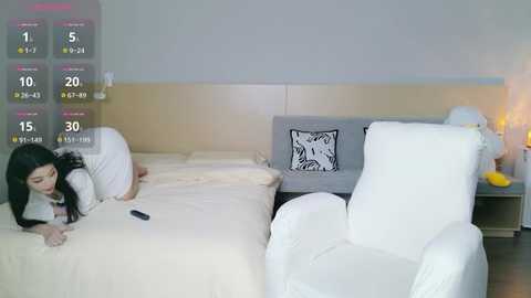 A video of an Asian woman with long black hair lying on a beige bed, wearing a white robe, in a modern bedroom with white armchairs and a gray headboard.