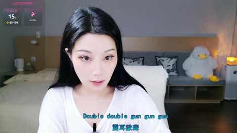 Video of an East Asian woman with long black hair and pale skin, wearing a white robe, sitting in a modern bedroom with a bed, lamp, and stuffed duck.