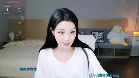 Video of an East Asian woman with long black hair, wearing a white t-shirt, sitting in a modern bedroom with a white bed, grey couch, and stuffed animal.