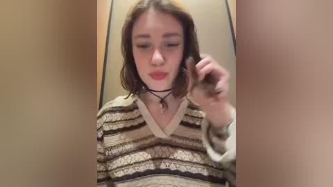 A video of a young woman with light skin and shoulder-length brown hair, wearing a patterned, beige sweater, holding a black phone in a public restroom stall.