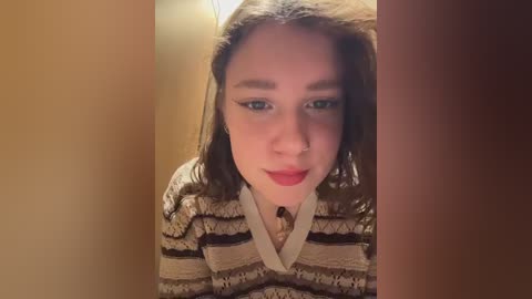 Video of a young woman with fair skin, dark brown hair, wearing a beige and brown striped sweater. She has a neutral expression and is looking directly at the camera.