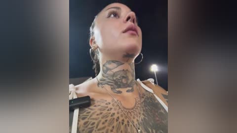 Media: Video of a woman with multiple tattoos, including a large one on her neck and chest, wearing hoop earrings, looking upward, in a dimly lit room with a spotlight.