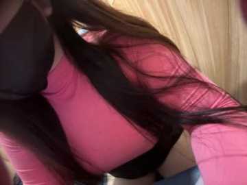 A close-up video of a woman's lower torso, showing a black bra and a pink top with sheer, mesh sleeves, her long, straight dark hair flowing around her.