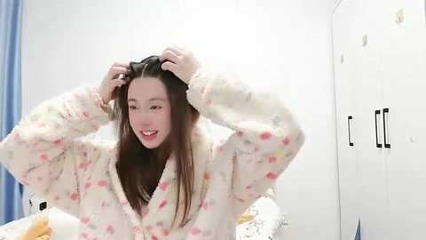 Video of an Asian woman with straight, shoulder-length brown hair, wearing a fluffy, light pink pajama set with pastel floral patterns, playfully adjusting her hair. She smiles warmly. Background includes a white wardrobe and a blue curtain.